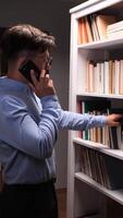 vertical Young male librarian searching for a book while talking on a smartphone, standing beside a white bookshelf in a library or bookstore, engaged in conversation and assisting a client video