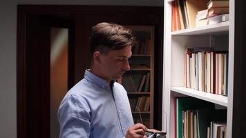 Young office worker cleaning eyeglasses with microfiber cloth, standing near bookshelves in library or home office, preparing for reading and working video