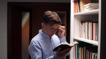Young man holding and reading a book while adjusting his glasses, standing next to a white bookshelf full of books, in a library or home study video