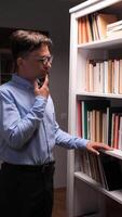 vertical A man in a library doesn't know which book to choose. The confused man shrugs his shoulders, doesn't know what to do. The man couldn't find the right book on the bookshelf. video