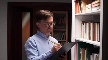 Young male librarian using tablet and consulting book catalog in library bookshelf, searching for specific book information, managing inventory, or assisting customers video
