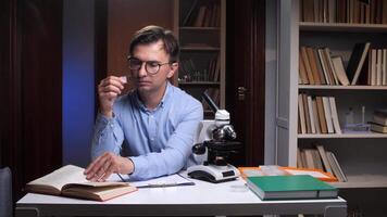A man in glasses sitting at a desk with a microscope and books video