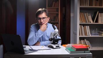 A man in glasses sitting at a desk with a laptop and microscope video