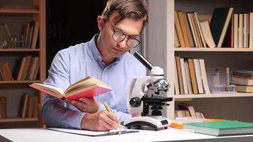 A man in glasses is writing on a book while looking at a microscope video