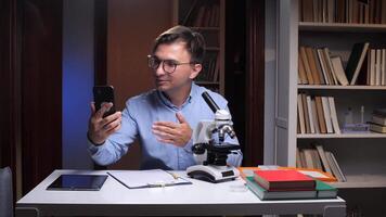 A man in glasses sitting at a desk with a microscope and a phone video