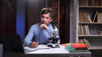 A man sitting at a desk with a microscope and laptop video