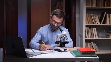 A man in glasses is writing on a piece of paper while looking at a microscope video