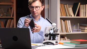 A man in glasses holding a cell phone and looking at a microscope video