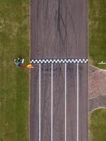 An aerial view of a race track featuring a checkered finish line, bordered by grass, with tire marks, equipment, and a person in an orange outfit visible. photo