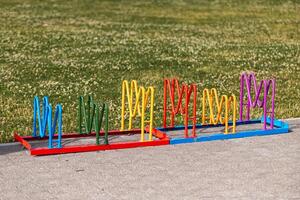 A vibrant bike rack with wavy loops in blue, green, yellow, red, and purple stands on a paved surface beside grass with small white flowers. photo
