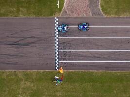 Two go karts cross a checkered finish line on a paved track, with a race official holding flags nearby. Tire marks and a brick walkway are visible. photo