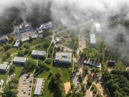 Cluster of modern buildings surrounded by trees and open green spaces, with pathways, parking lots, and light fog partially obscuring the scene. photo