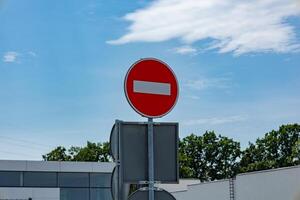A bright red no entry sign is visible against a clear blue sky, surrounded by greenery and a modern building in the background photo