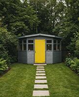 Modern grey garden shed with bright yellow door stands on green lawn, surrounded by trees and greenery. Shed has flat roof and windows. Path of square stones leads to the entrance. Ideal for adu use. photo