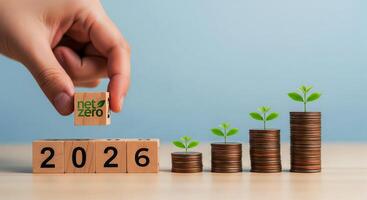 Hand places net zero cube onto wooden blocks showing the year 2026 with growing money plants photo