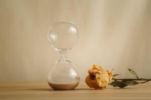 A simple hourglass on a wooden table with dry rose a beige backdrop, symbolizing time flow, aging, and minimalistic living. Represents timeless themes in a creative presentation for stylish concepts. photo