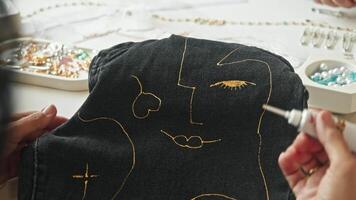A woman creates a unique design on dark denim fabric, using golden paints to depict a portrait. video