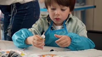 A little boy is using glue while crafting denim clothes and doing needlework in a lesson. He is wearing apron and painting his jeans with paint. Portrait. video
