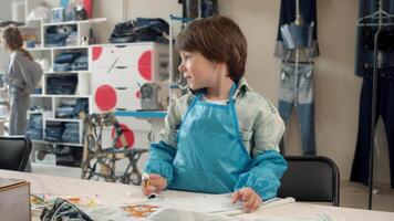 little boy is engaged in process of making handmade garments during an art class. He is wearing a protective apron and is using paint to decorate his jeans. video