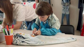 Cute little boy is creating craft clothes during art lesson or craft class. He is wearing a protective apron and is using paint to decorate his jeans. video