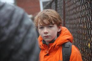 A young boy wearing an orange jacket standing next to a fence photo