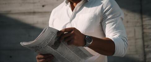 The man casually reading a newspaper outdoors while checking the time on his watch. photo