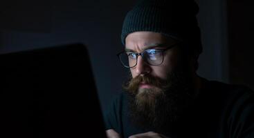 A man with a beard and glasses is looking at a laptop photo