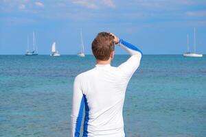 Rear view of a man standing on the beach in a sun protected shirt, looking at the sea and sailboats. Summer vacation and water sports concept photo