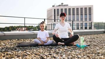 Mother and daughter performing yoga meditation on rooftop during sunny day. Family bonding through relaxation and mindfulness practice in outdoor urban environment. video