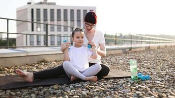 Mother sitting with daughter on rooftop, practicing fitness exercises using small dumbbells, enjoying bonding time. Child learning proper exercise techniques from parent. video