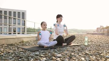Mother and daughter sitting outdoors on rooftop doing exercises with dumbbells during sunrise. Both models smiling wearing casual sportswear promoting family fitness, outdoor activity video