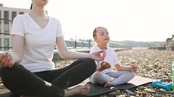 Mother sits on yoga mat practicing meditation with young daughter, both in lotus position enjoying calm morning ambiance following healthy lifestyle routine video