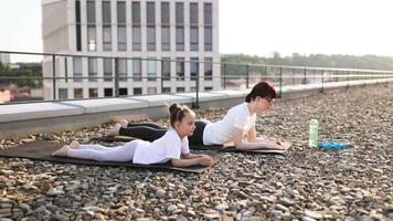 Mother and daughter engaging in plank exercises on rooftop space highlighting fitness and bonding. Bright day atmosphere emphasizing outdoor workout. video