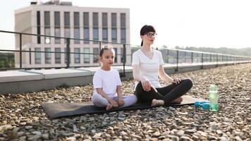 Mother and daughter practicing yoga together on rooftop. Urban scene emphasizes relaxation and healthy lifestyle. Both sitting cross legged in meditative posture, showcasing family bonding video