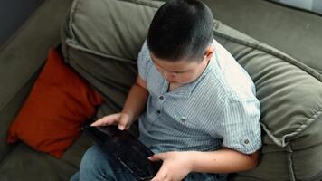 boy sits on olive couch with digital tablet in blue striped shirt, indoor relaxed atmosphere, concept of digital education, online gaming, home technology, top view video