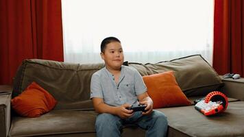 smiling Asian boy holds game controller on sofa, striped shirt and jeans, focused on game play, modern casual home interior, cozy green couch with orange pillow video