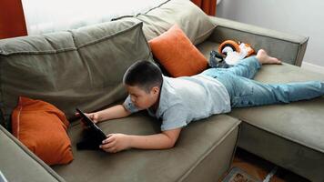 boy lying on olive couch with digital tablet in blue striped shirt, indoor relaxed atmosphere, concept of digital education, online gaming, home technology video