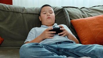 Asian boy holds game controller on sofa, striped shirt and jeans, focused on game play, modern casual home interior, cozy green couch with orange pillow, sweating of the palms video