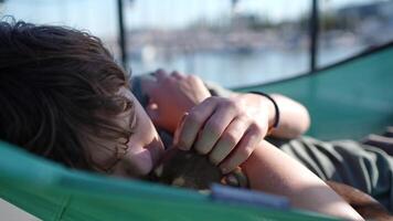 Teenage boy lying peacefully in green hammock, gently stroking small brown chihuahua near marina waterfront with sailboats and soft afternoon sunlight video