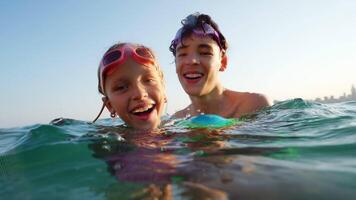 Cheerful siblings wearing swimming goggles enjoying playful moments with colorful inflatable ball, splashing and laughing while experiencing ocean vacation under bright sunlight video