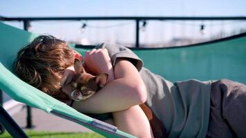 Young boy peacefully resting on rooftop deck chair, tenderly hugging small chihuahua while enjoying warm sunlight and urban skyline backdrop video