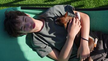 Top view of a peaceful teenager resting with tiny chihuahua companion, sharing tranquil summer moment on garden camping cot, embodying serene bond of friendship and comfort video