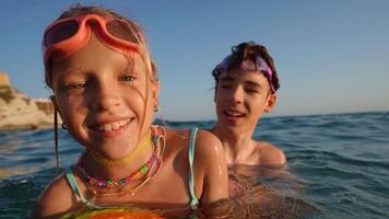 Brother and sister splashing playfully in sparkling ocean waters, wearing vibrant swimming goggles and colorful necklaces during summer vacation moments of pure childhood joy video