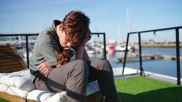 Teenager boy sitting on a white deckchair is embracing his small chihuahua dog in a marina, with yachts and sailboats moored in the background video