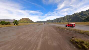 Cinematic FPV drone shot of red jeeps driving across volcanic black sand plains near Mount Bromo, with lush mountain backdrop in East Java, Indonesia. Concept of travel, tour, and adventure. video