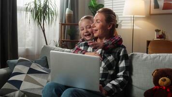 Happy joyful Caucasian child kid girl daughter hugging mother working on laptop at home jumping behind sofa embracing smiling talking enjoying family parenting work life balance freelance living room video