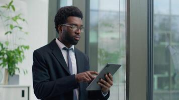 Thoughtful African American businessman in office concentrate using digital tablet looking out window focused business male man guy using technology device thinking planning strategy professional work video