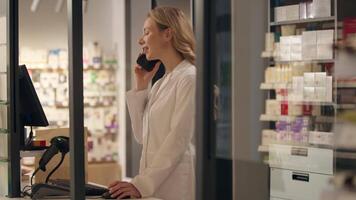 Pharmacy worker Caucasian woman in medical coat talk mobile phone looking at computer check drugs pills stock medicine inventory drugstore health care products professional consultation cellphone call video