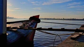 A traditional wooden boat docked by a dock, with the sun rising over the water in the background. video