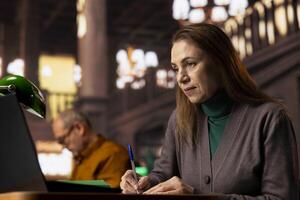 Elderly academic woman prepares documents for her university coursework, transcribing from literature and textbooks. Student focused on research and education in a quiet public library. photo